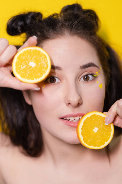 肌の近くにオレンジの果実のスライスを持つ若いブルネットの女性黄色の背景に顔。おいしいジューシーな果物。熱帯果実、健康食品。柑橘類のスライス。おいしいジューシーなフルーツ - 写真・画像