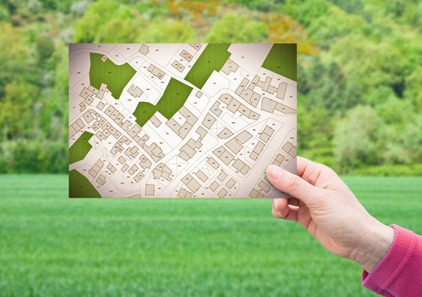 Διαχείριση οικοπέδου - έννοια της ακίνητης περιουσίας με μια κενή γη σε μια πράσινη κατοικημένη περιοχή διαθέσιμη για κατασκευή κτιρίου με το χέρι κρατώντας μια κάρτα για ένα φανταστικό κτηματολογικό χάρτη - Φωτογραφία, εικόνα