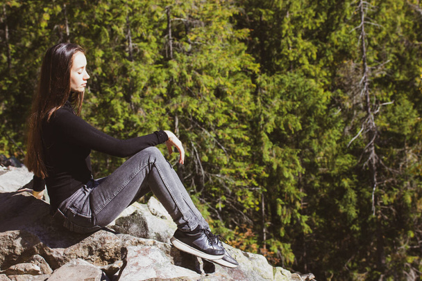 Krásná bílá dívka s dlouhými tmavými vlasy sedí na vrcholu hory v zeleném jehličnatém lese, prostor pro text. Mladá žena, sama v lese. Dobrodružství, výlet. Užívám si cestování po přírodě. - Fotografie, Obrázek