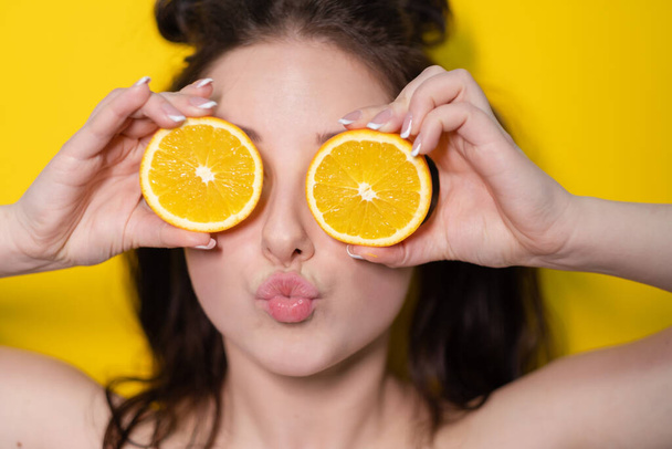 jong brunette vrouw meisje met slice van oranje fruit in de buurt van huid gezicht op gele achtergrond. lekker sappig fruit. Tropische vruchten, gezond voedsel. citrusschijfje. smakelijk sappig fruit - Foto, afbeelding