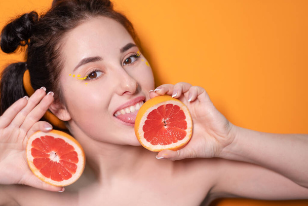 sladká radostná brunetka žena dívka fena s ostříhaným plátek grapefruitu v blízkosti kůže tvář na oranžovém pozadí. chutné šťavnaté ovoce. Tropické citrusové plody, zdravé jídlo. úbytek hmotnosti - Fotografie, Obrázek