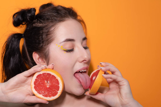 甘い喜びのブルネットの女性オレンジの背景に肌の近くにグレープフルーツの切り身のスライスを持つ女性。おいしいジューシーな果物。熱帯柑橘類、健康食品。体重減少 - 写真・画像