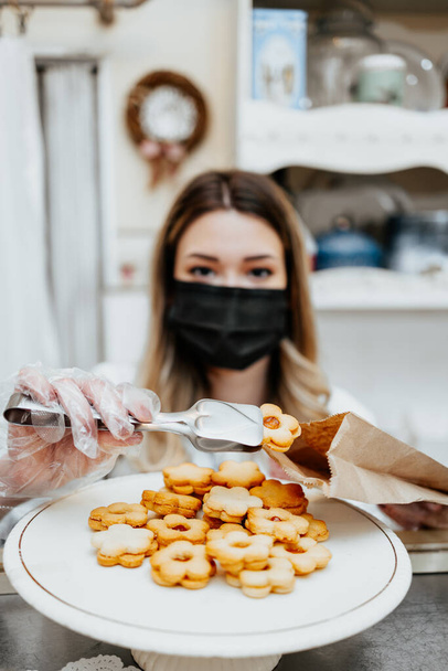 Beautiful young female seller with protective face mask working in a confectionery shop or bakery. She is selling and packing delicious sweets. Coronavirus and small business concept. - Фото, зображення