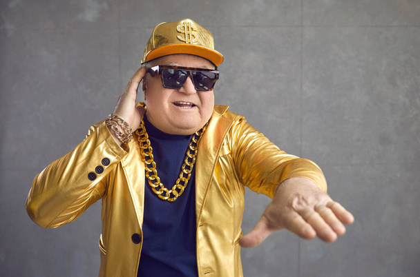 Funny senior man in golden jacket, baseball cap and chain necklace playing music at disco party - Fotografie, Obrázek