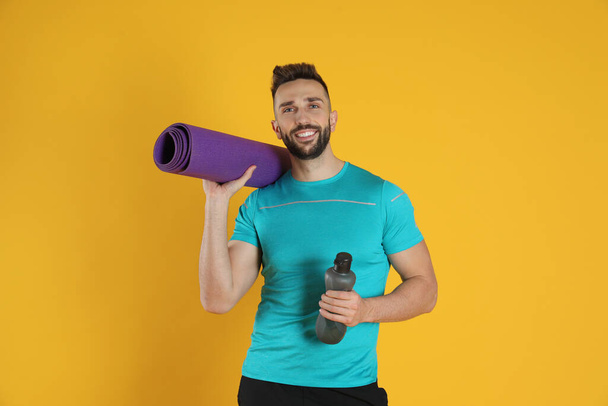 Handsome man with yoga mat and bottle of water on yellow background - Φωτογραφία, εικόνα