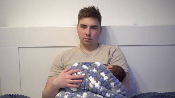 疲れ切った若いお父さんは子供をベッドの上で抱きしめる。腕の中に新生児を持つ熱狂的な疲れの表情を持つ男. - 写真・画像