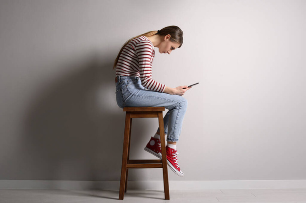 Woman with bad posture using tablet while sitting on stool near light grey wall indoors - Foto, imagen