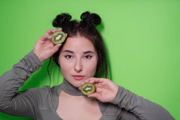 Krásný portrét atraktivní mladé ženy s čistou zářivou pletí tvář s kiwi půlky, brunetka dívka s holými rameny drží kiwi na zeleném pozadí. Tropické ovoce, zdravé jídlo - Fotografie, Obrázek