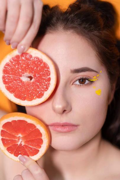 сладкая радостная брюнетка женщина женщина с обрезанным ломтиком грейпфрута возле лица кожи на оранжевом фоне. вкусные сочные фрукты. Тропические цитрусовые, здоровая пища. потеря веса - Фото, изображение