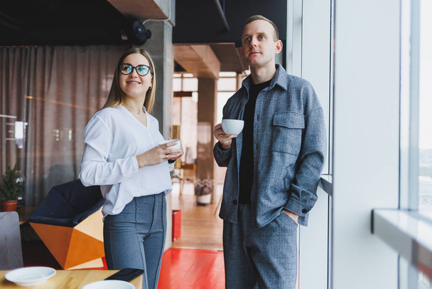 Ένας επιχειρηματίας και μια επιχειρηματίας πίνουν καφέ μαζί, στέκονται μπροστά από τα παράθυρα ενός κτιρίου γραφείων με θέα την πόλη. Δύο επιχειρηματίες με αυτοπεποίθηση εργάζονται σε ένα σύγχρονο γραφείο - Φωτογραφία, εικόνα