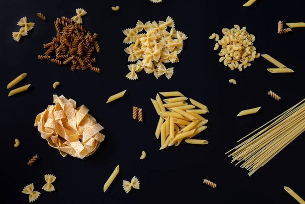 Different types of uncooked dried pasta on the black background. Raw pappardelle, penne, spaghetti, farfalle and fusilli. Italian pasta cooking concept. - Photo, Image