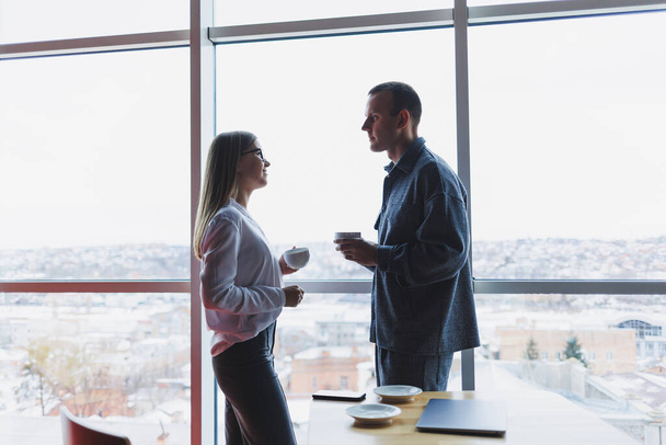 Business people stand relaxed during a work break with a cup of coffee in a modern office surrounded by glass overlooking a high-rise building. - Photo, Image