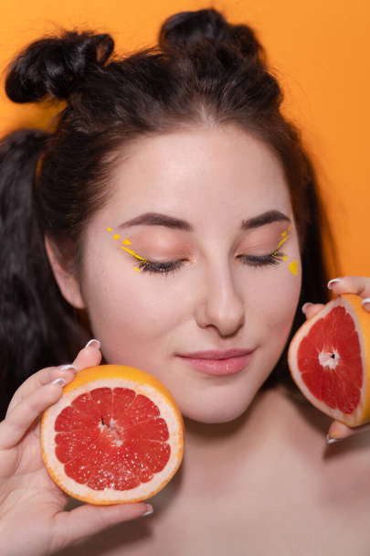甘い喜びのブルネットの女性オレンジの背景に肌の近くにグレープフルーツの切り身のスライスを持つ女性。おいしいジューシーな果物。熱帯柑橘類、健康食品。体重減少. - 写真・画像