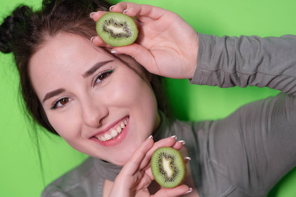 Schönheitsporträt einer attraktiven jungen Frau mit sauberem strahlenden Gesicht mit Kiwi-Hälften, brünettes Mädchen mit nackten Schultern, das Kiwi auf grünem Hintergrund hält. Tropische Früchte, gesunde Ernährung. - Foto, Bild
