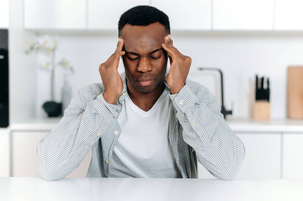 Sad african american guy in stress, sitting at the table alone, in casual clothes, experiencing fatigue, headache, migraine, massaged his temples, closes his eyes, needs rest, medicines and treatment. - Photo, image
