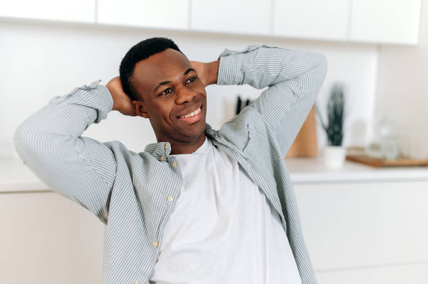 Portret przystojnego Afroamerykanina. Czarny atrakcyjny facet ubrany w stylowe ubrania casual siedzi w domu w salonie w spokojnej pozie z rękami za głową, uśmiechając się szczęśliwie - Zdjęcie, obraz