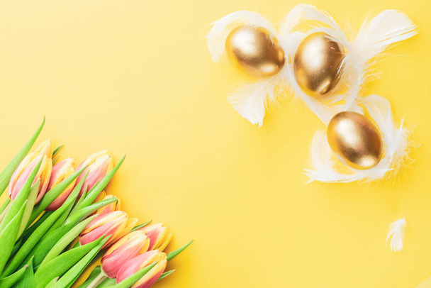 Cesta de ovos de Páscoa. Ovo dourado em cesta com tulipas de primavera, penas brancas em fundo amarelo pastel em decoração Feliz Páscoa. Decoração tradicional à luz do sol. Vista superior - Foto, Imagem