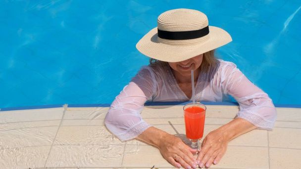mosolygó szőke nő szalmakalapban ellazul és narancssárga koktélt iszik a medence mellett egy napos nyári napon. Nyári hangulat koncepció, nyaralás, all inclusive - Fotó, kép