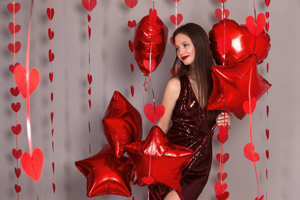 Ένα νεαρό κορίτσι που κινηματογραφεί στο στούντιο σε γκρι φόντο με μπαλόνια κόκκινης καρδιάς, οι διακοπές είναι Ημέρα του Αγίου Βαλεντίνου. Γυναικεία χέρια που κρατάνε κόκκινες καρδιές κοιτάζοντας από κοντά την κάμερα. - Φωτογραφία, εικόνα