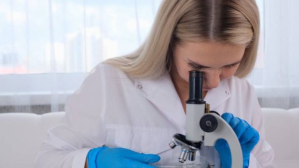 Asistente de laboratorio femenino analiza muestras utilizando un microscopio mientras trabaja en una mesa en un laboratorio, haciendo investigación - Foto, Imagen