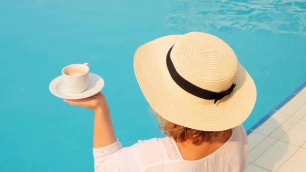 Femme de 50 ans qui prend son petit déjeuner au bord de la piscine avec un chapeau de paille et une robe blanche. femme assise près de la piscine avec une tasse de café. Bonjour et planification du jour - Séquence, vidéo