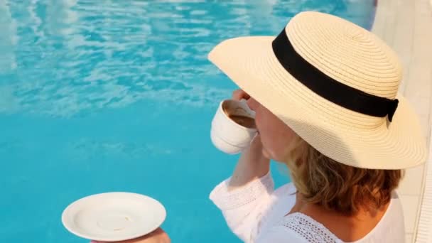 Κοντινό πλάνο μιας γυναίκας ηλικίας 50-55 ετών σε ένα ψάθινο καπέλο πίνοντας από ένα φλιτζάνι καφέ δίπλα σε μια μπλε πισίνα, πολυτελή καλημέρα, έναρξη της ημέρας - Πλάνα, βίντεο