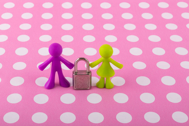 Una serratura in miniatura si trova tra le figure simboliche di un uomo e una donna su uno sfondo rosa con piselli bianchi - Foto, immagini