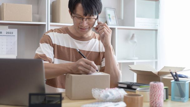 Asyalı adam siparişlerini doğrulamak için bir telefondan online sipariş alıyor ve müşterilerle sohbet ediyor, ürünleri internette satıyor veya evde serbest iş yapıyor, evde çalışıyor ve iş sahibi oluyor.. - Fotoğraf, Görsel