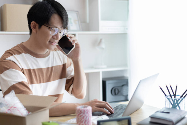Hombre asiático está tomando pedidos en línea desde un teléfono y charlar con los clientes para confirmar su pedido, la venta de productos en línea o haciendo trabajo independiente en casa, Trabajar en casa y ser dueño de negocios. - Foto, imagen
