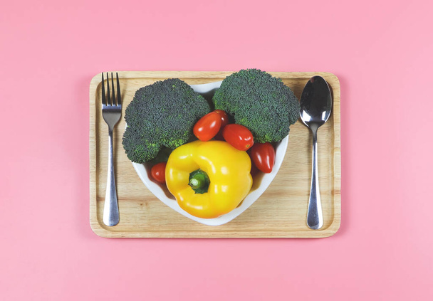 Bovenaanzicht of platte lay van groenten capsicum, broccoli en tomaat in hartvorm plaat met vork en lepel in houten lade op roze achtergrond met kopieerruimte, gezonde levensstijl en gewichtsverlies concept. - Foto, afbeelding