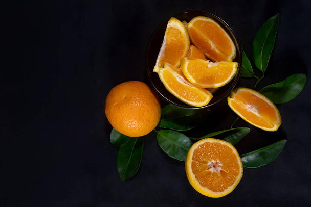 Rebanadas y cuñas de naranjas frescas con hojas naranjas y verdes sobre un fondo negro texturizado con vista desde arriba hacia abajo. - Foto, Imagen