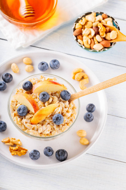 Oatmeal with blueberries and sliced apple in bowl. Oatmeal porridge with berries, nuts and honey on a white boards. Healthy breakfast concept. Top view - Foto, Bild