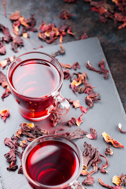 Red hibiscus tea in glass mug on a dark background. Cup of hibiscus tea and dry hibiscus petals on a slate board. Copy space - Foto, Imagen