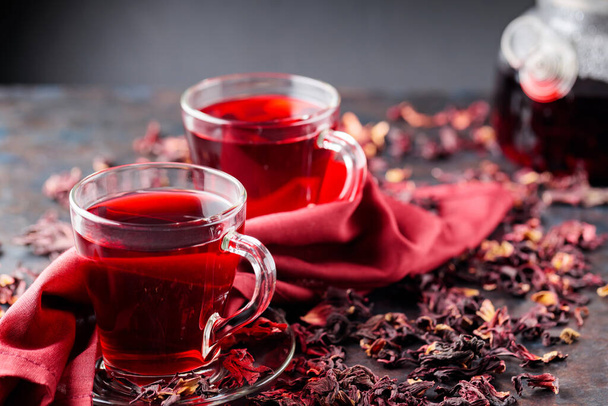 Hibiscus thee in glazen beker en een bordeaux servet. Kopje hibiscus thee en droge hibiscus bloemblaadjes op een donkere achtergrond. Gezonde natuurlijke afslanken drank - Foto, afbeelding