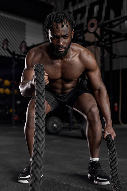 Young black man doing strength training using battle ropes at modern dark gym. - Foto, afbeelding