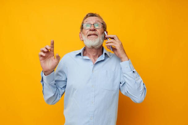 Senior grey-haired man in a blue shirt and glasses talking on the phone isolated background - Фото, изображение