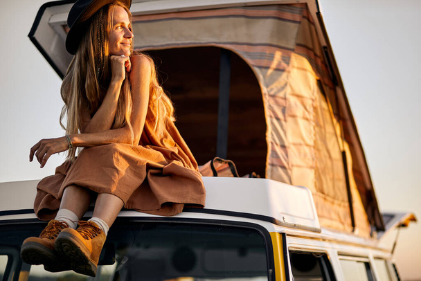 Atractiva dama encantadora está en el techo de mini furgoneta amarilla, disfrutar de la puesta de sol desde la parte superior - Foto, imagen