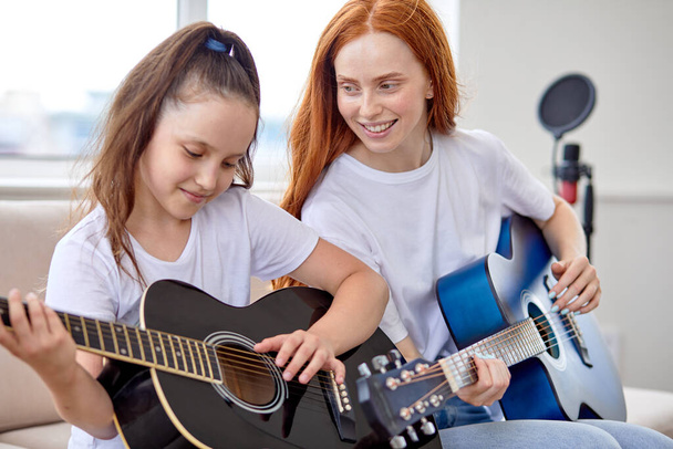Pretty redhead adult woman and little girl at home family time sitting on sofa playing guitar - Foto, immagini