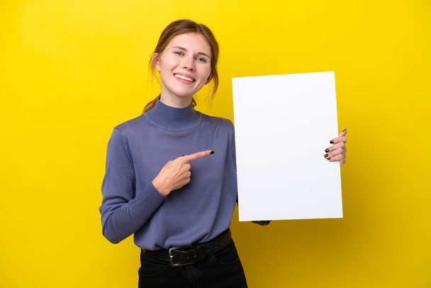 若い英語の女性は幸せな表情で空のプラカードを保持し、それを指す黄色の背景に隔離 - 写真・画像