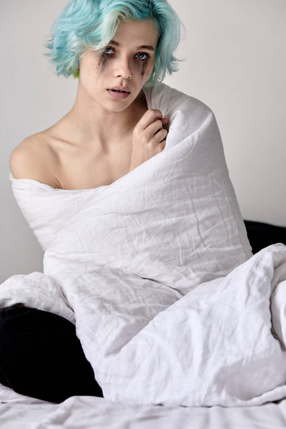 Worried young female sitting on bed wrapped in blanket. Stressed lady at home - 写真・画像