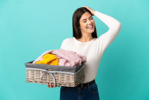 Teenager Brazilian girl holding a clothes basket isolated on blue background smiling a lot - Фото, изображение