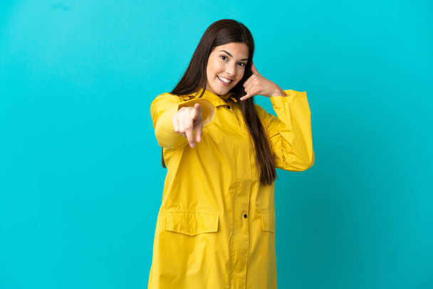 Adolescente menina brasileira vestindo um casaco à prova de chuva sobre fundo azul isolado fazendo gesto telefone e apontando frente - Foto, Imagem