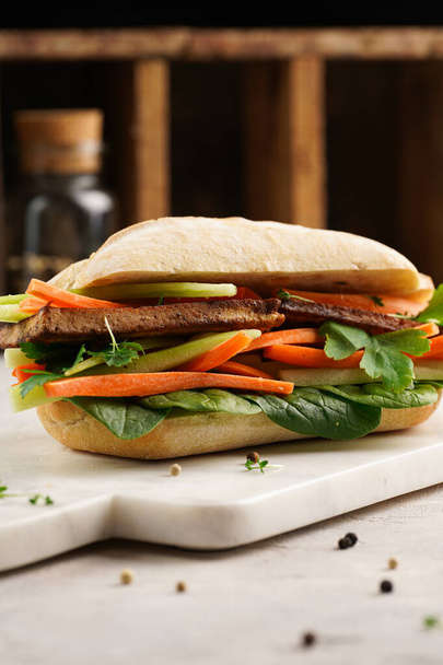 Traditional vietnamese banh mi sandwich with sliced smoked tofu, fresh spinach, shredded carrots and peeled cucumbers, cilantro on marble board on light grey textured background - Foto, afbeelding