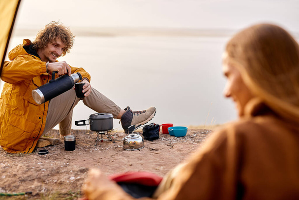 Smiling curly man puring tea into cup from thermos, smiling, enjoy travel - Foto, Imagen