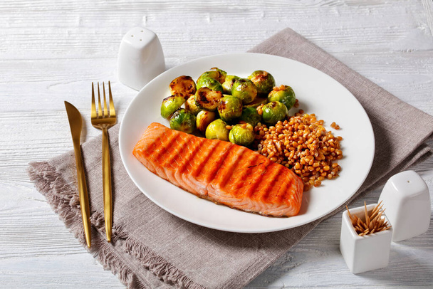 grilled salmon fillet, roast brussel sprouts and boiled farro on a white plate on a wooden table, horizontal view from above - Zdjęcie, obraz