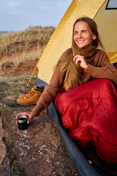 woman in sweater drinking tea while sitting in tent, travelling camping in wild nature - 写真・画像