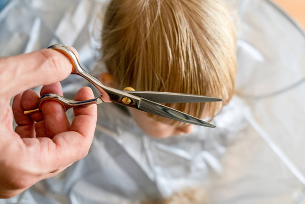 Les mains de l'homme faisant blonde tout-petit coupe de cheveux avec des ciseaux de coiffeur à la maison. Bébé garçon coupe de cheveux bricolage - Photo, image