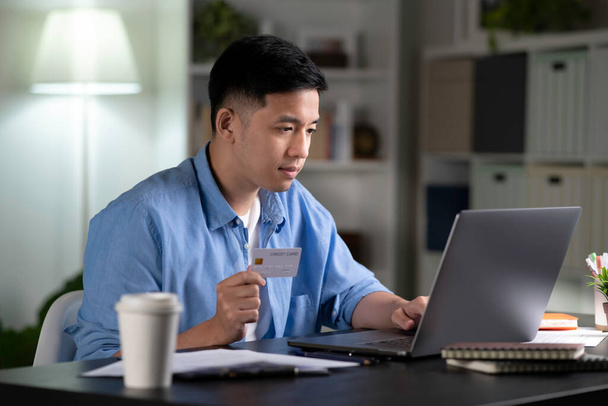 Magabiztos fiatal ázsiai férfi, online vásárló online vásárlás, böngésző bolt weboldalak, hogy az online fizetési tranzakció a laptop és tartja hitelkártya otthon. - Fotó, kép