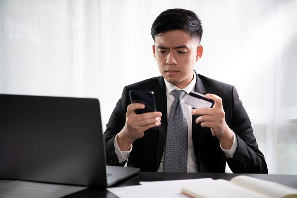 Nuori aasialainen liikemies virallisessa puku istuu kotona toimistossa kannettava tietokone näyttää onneton harmitti ja turhautunut, kun käytät luottokorttia ja älypuhelin. Liikemies näköinen stressaantunut ja tyytymätön tehdessään epäonnistunut digitaalinen online-maksu usi - Valokuva, kuva