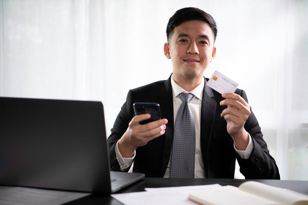 Inteligentny młody azjatycki biznesmen siedzi w biurze z laptopem uśmiechnięty i szczęśliwy podczas korzystania z karty kredytowej z aplikacji mobilnej zakupów online na smartfonie. Młody azjatycki przedsiębiorca wygląda pewnie i zadowolony z cyfrowej płatności online tra - Zdjęcie, obraz